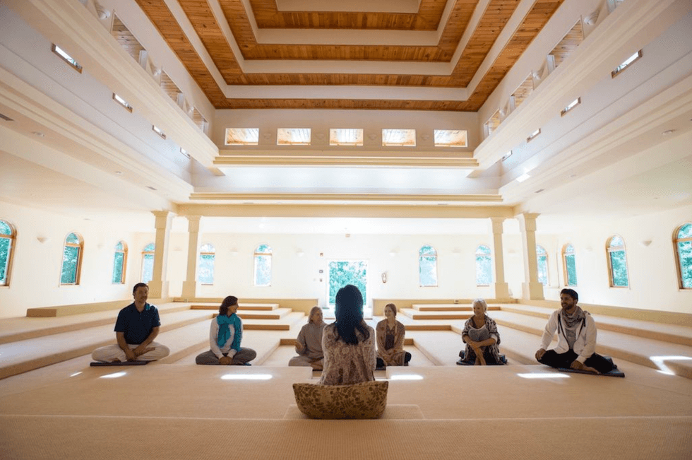 Group meditating in a bright, spacious room with tiered seating and wooden ceiling.
