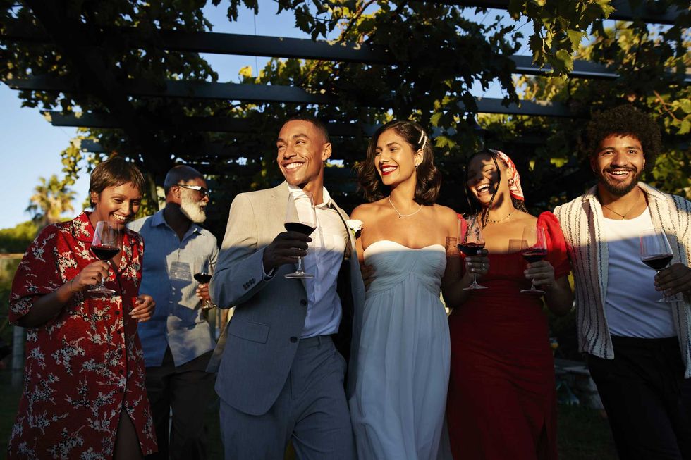 Group of people at an outdoor party, smiling and holding wine glasses under vine-covered pergola.