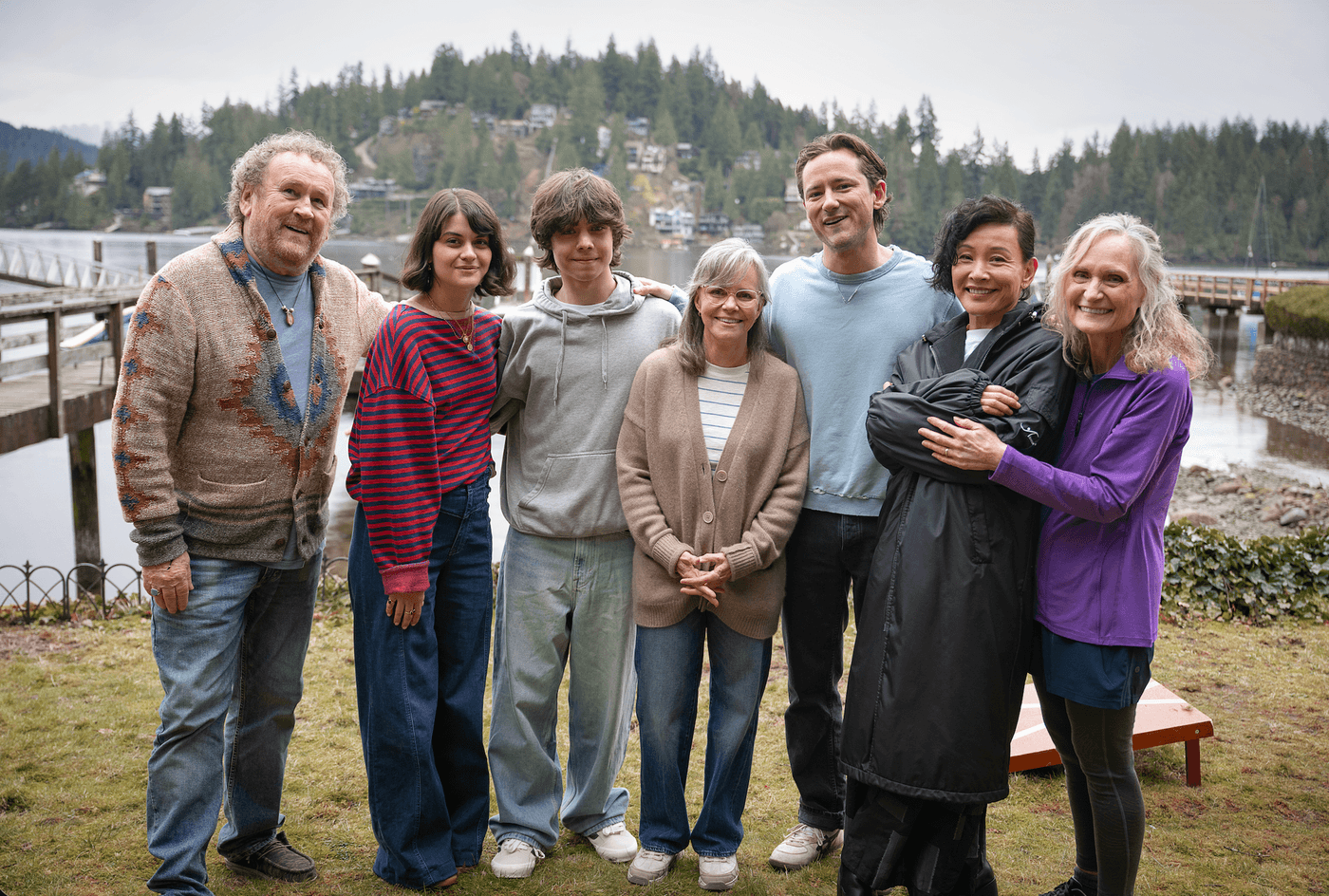 Group of seven people smiling by a lake, with a forested background.