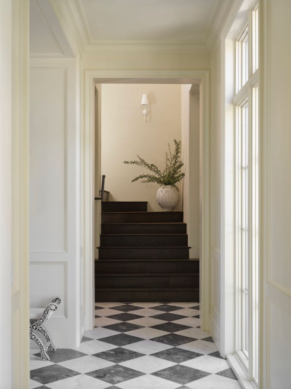 Hallway with checkered floor, dark steps, potted plant, and wall light.