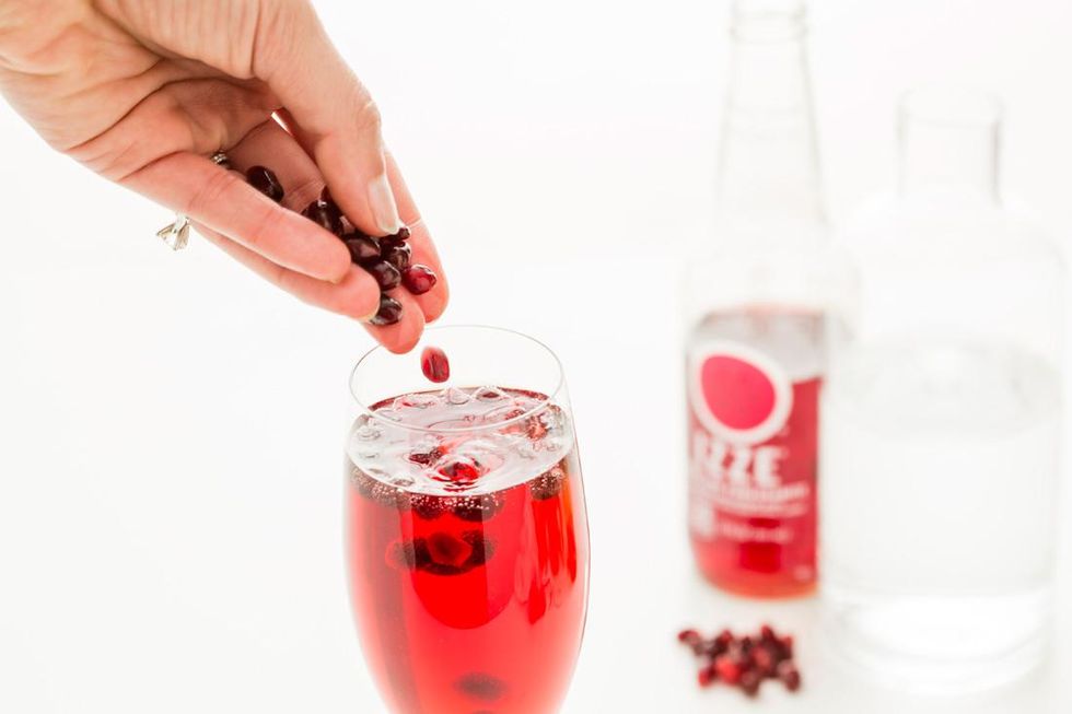 Hand dropping pomegranate seeds into a red drink with bottles in the background.