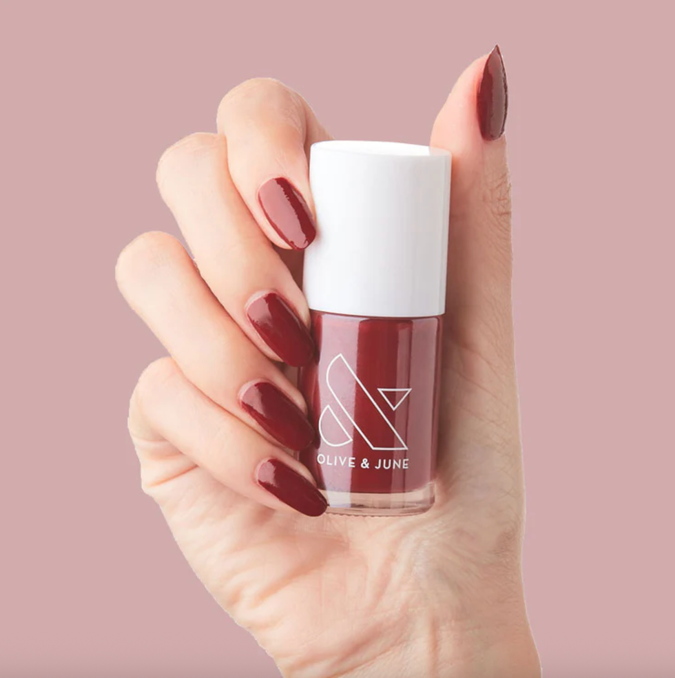 Hand holding a bottle of dark red nail polish against a pink background.