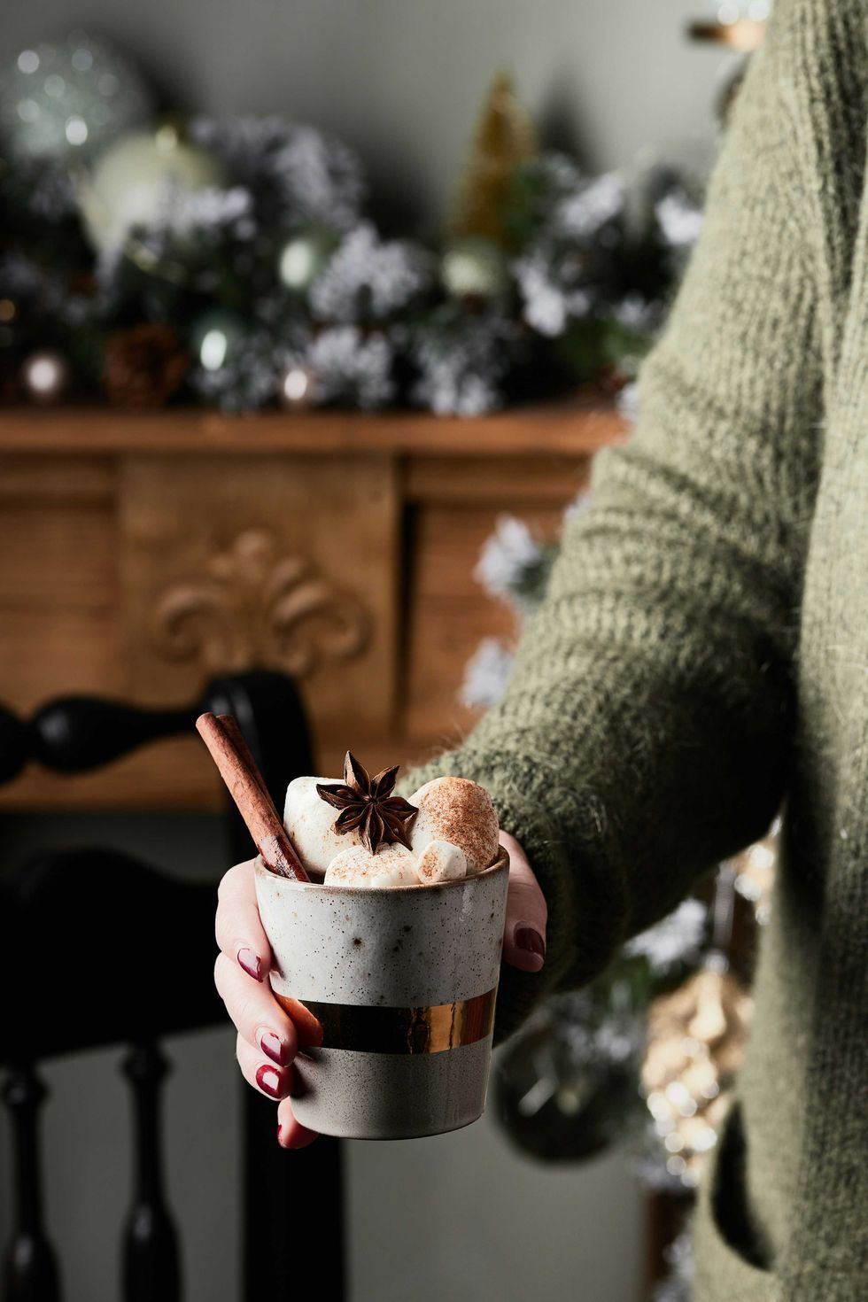Hand holding a festive cup with hot cocoa, marshmallows, cinnamon, and star anise.