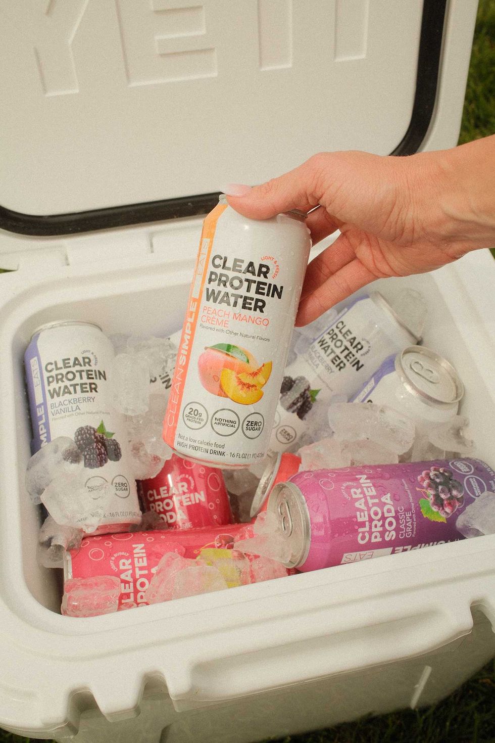 Hand holding a Peach Mango Clear Protein Water can from a cooler filled with assorted cans and ice.