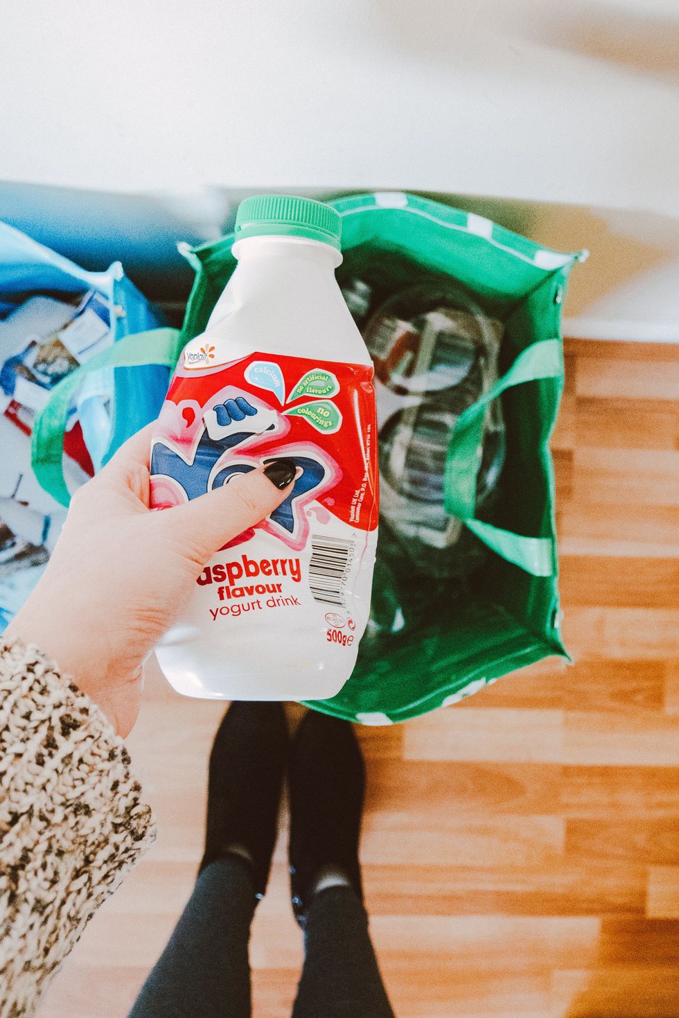 Hand holding raspberry yogurt drink above a shopping bag on wooden floor.
