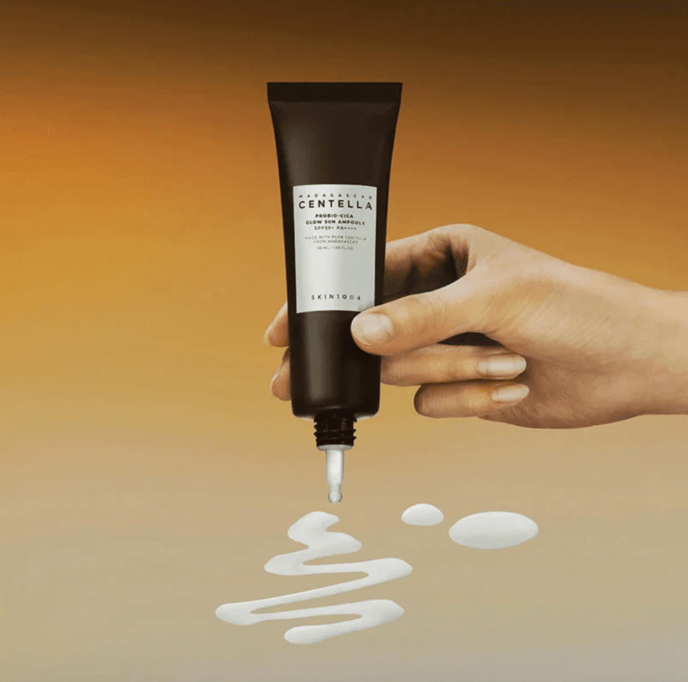 Hand holding SKIN1004 cream tube, dispensing white cream on gradient background.