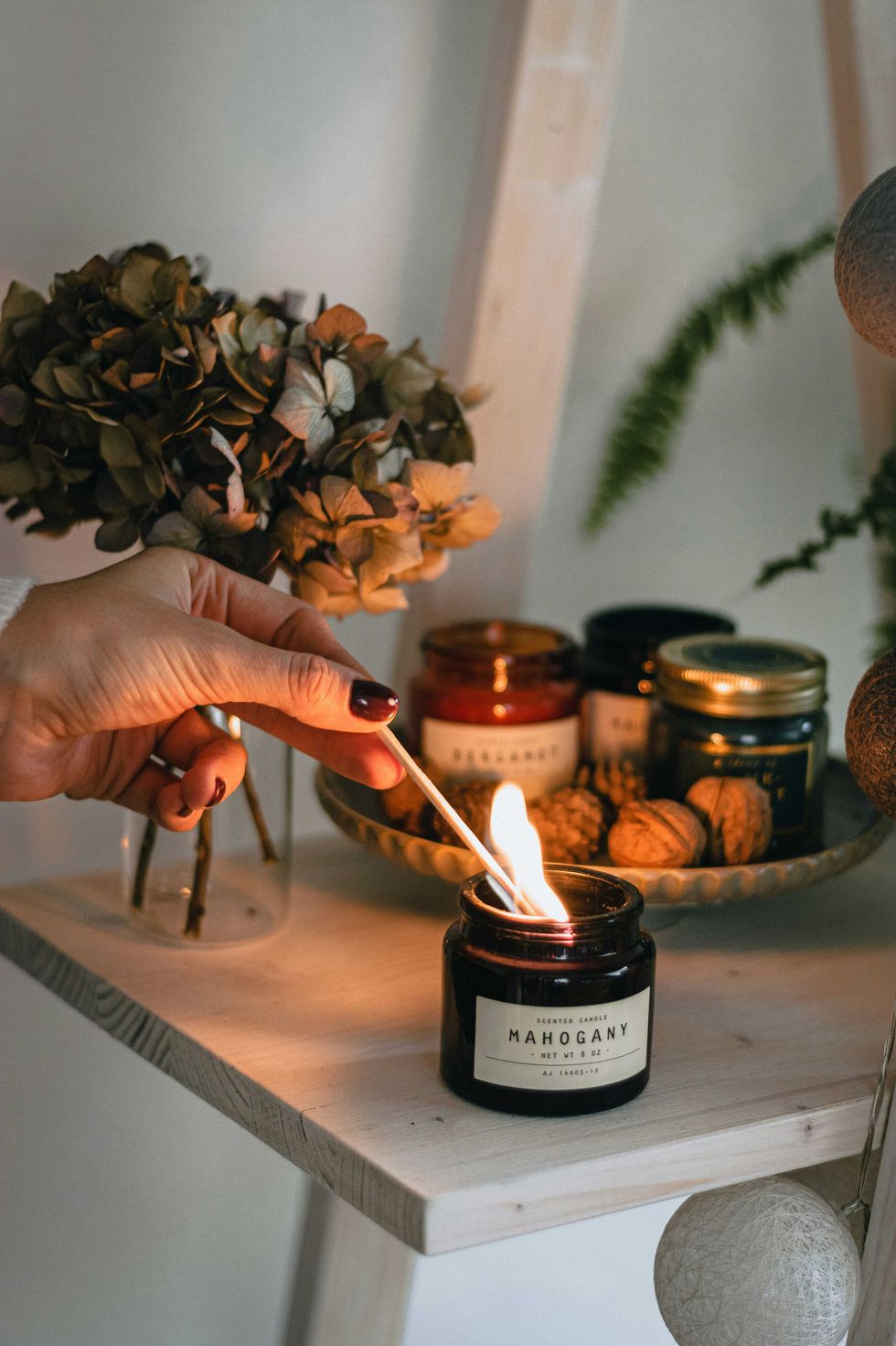 Hand lighting a mahogany-scented candle on a wooden shelf with dried flowers.