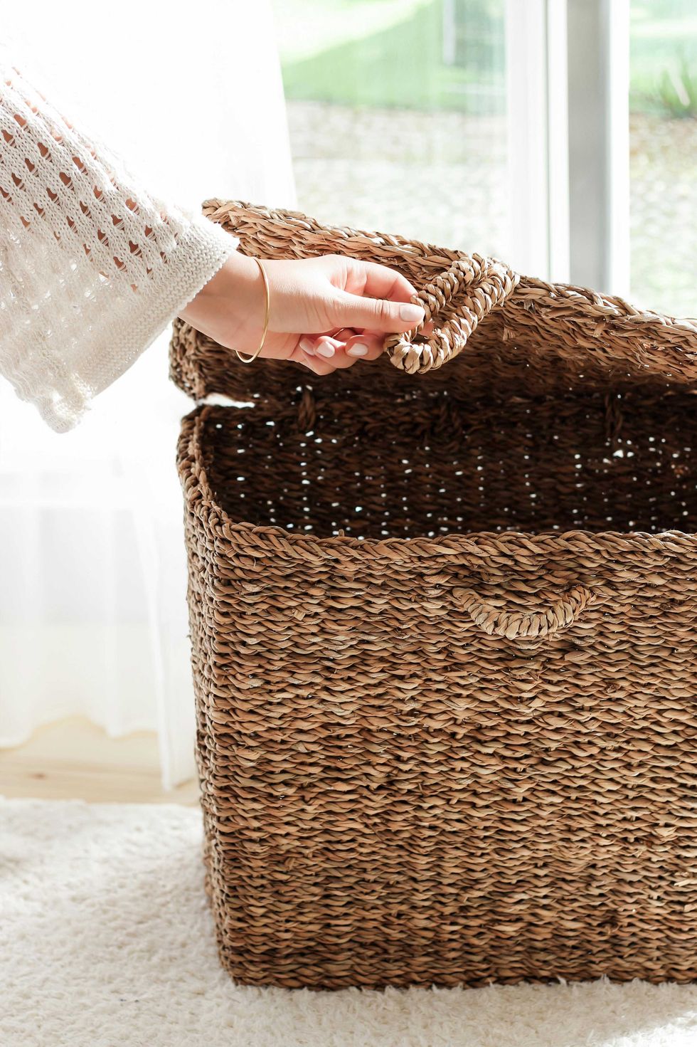 Hand opening the lid of a woven wicker basket near a bright window.