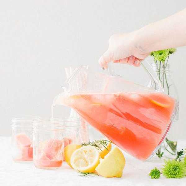 Hand pouring pink lemonade from a pitcher into three jars with lemons and flowers.