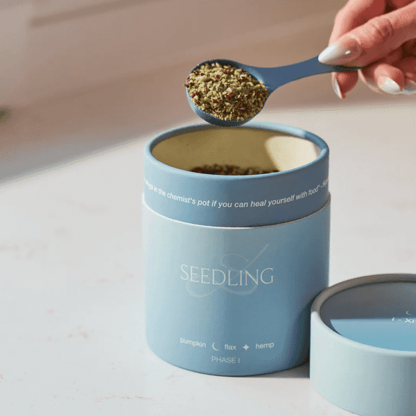 Hand scooping seeds from a blue "Seedling" container on a light surface.