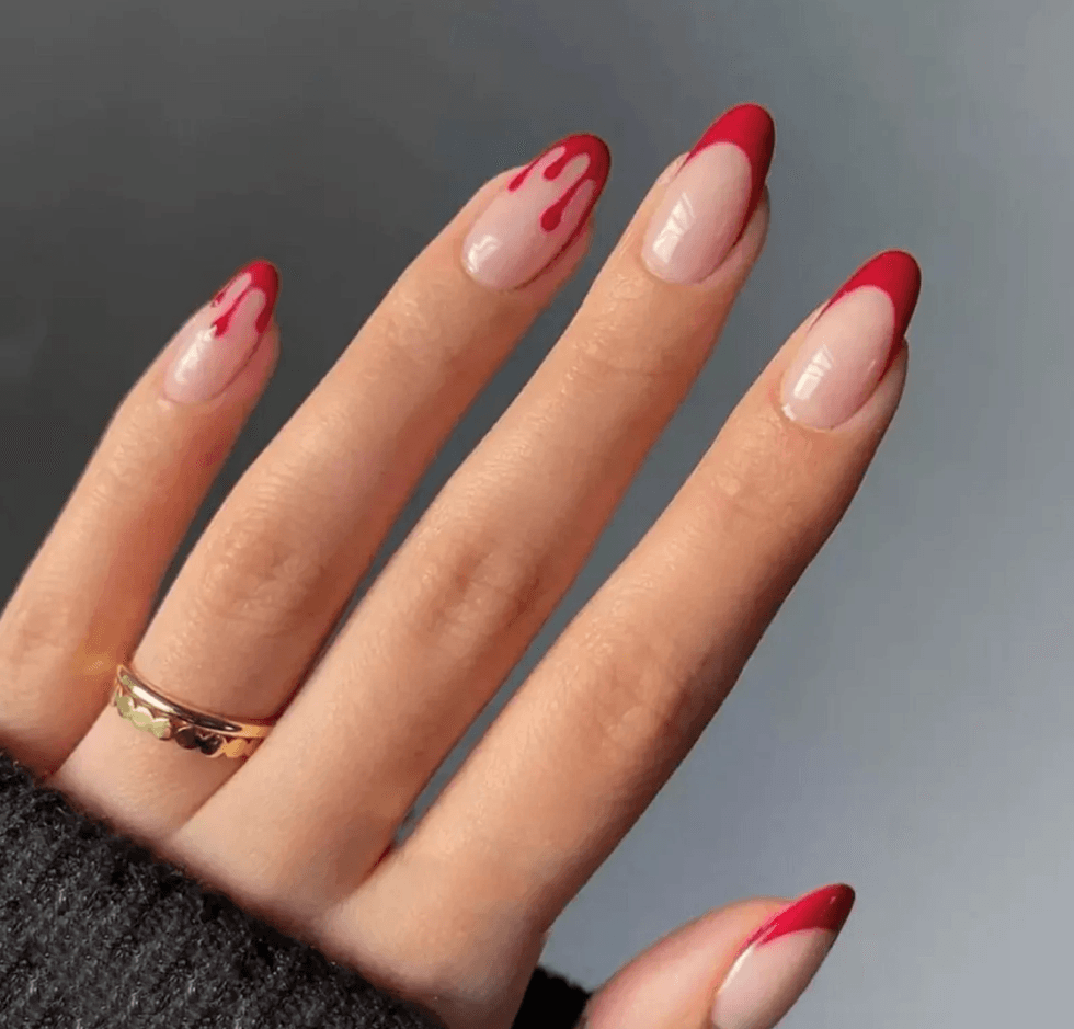 Hand with red drip nail art and gold ring on a gray background.
