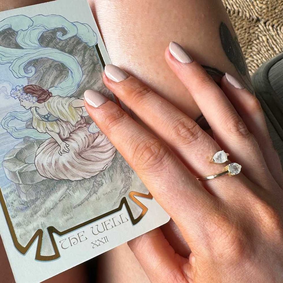 Hand with ring rests on leg holding "The Well" tarot card, showcasing elegant nails.