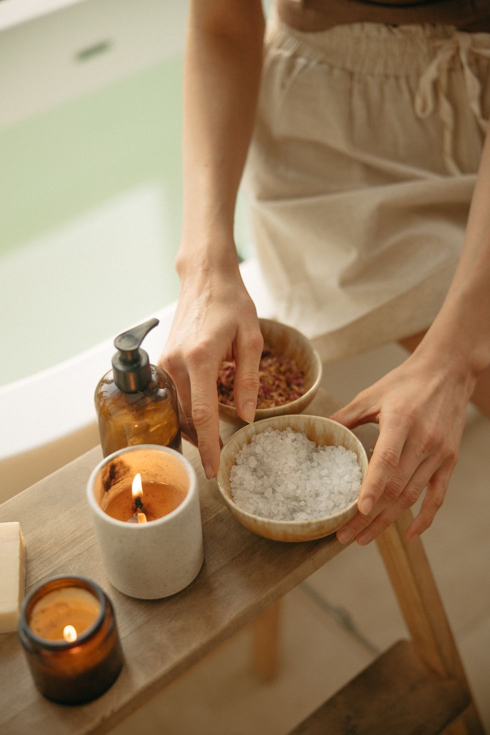 Hands arranging bath salts, candles, and a soap pump on a wooden surface by a tub.