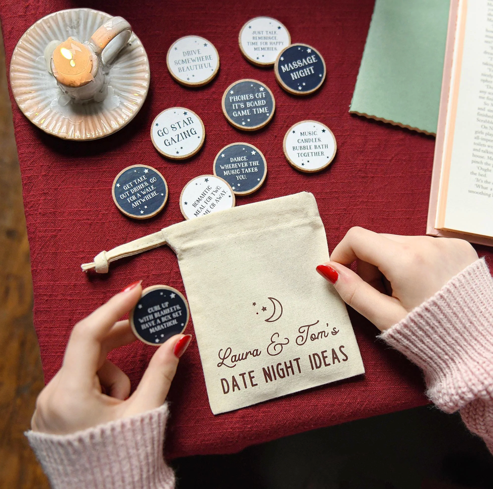 Hands holding a "Date Night Ideas" pouch with suggestion tokens on a red tablecloth.