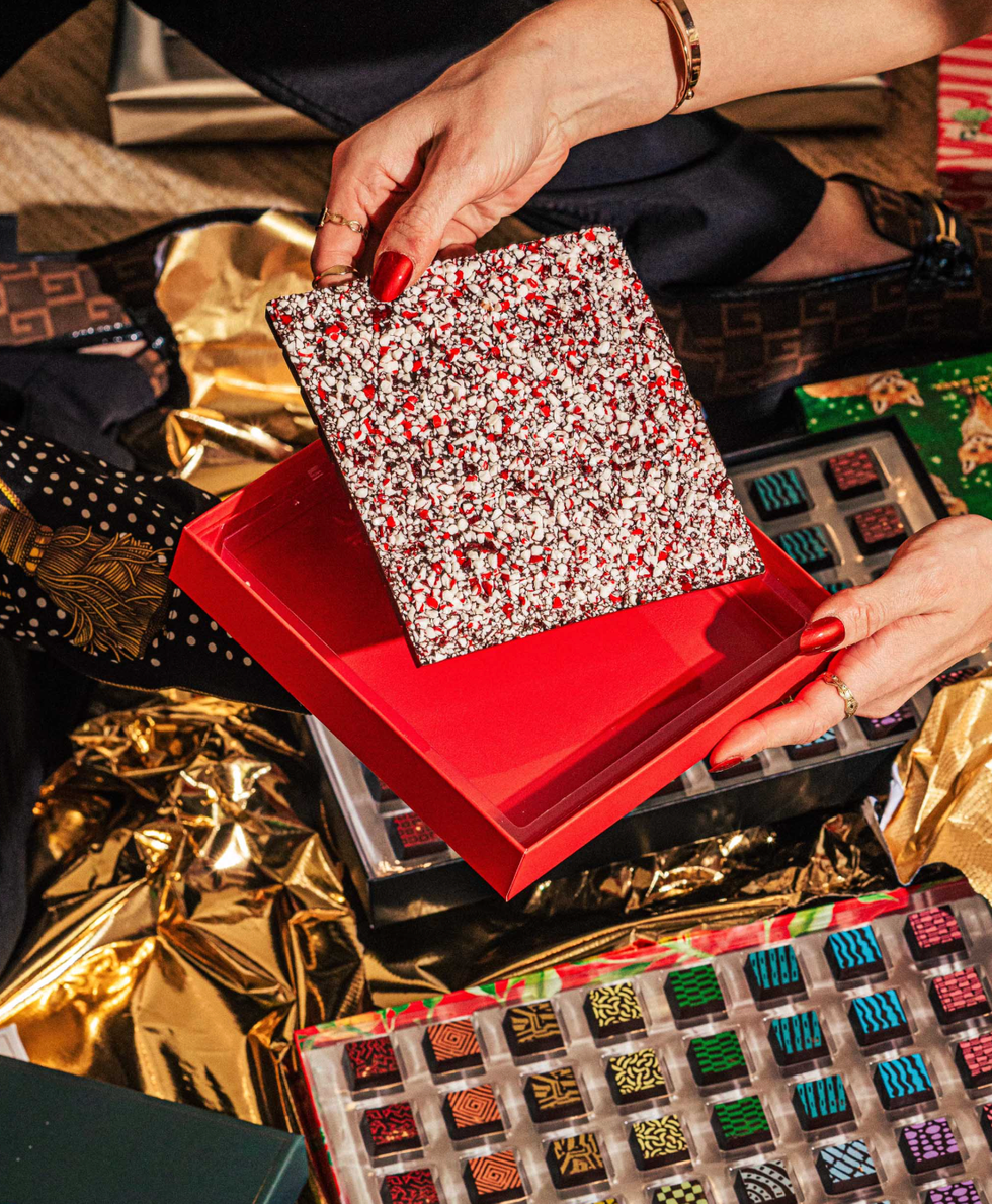 Hands holding a speckled chocolate slab over a red box, surrounded by assorted chocolates.