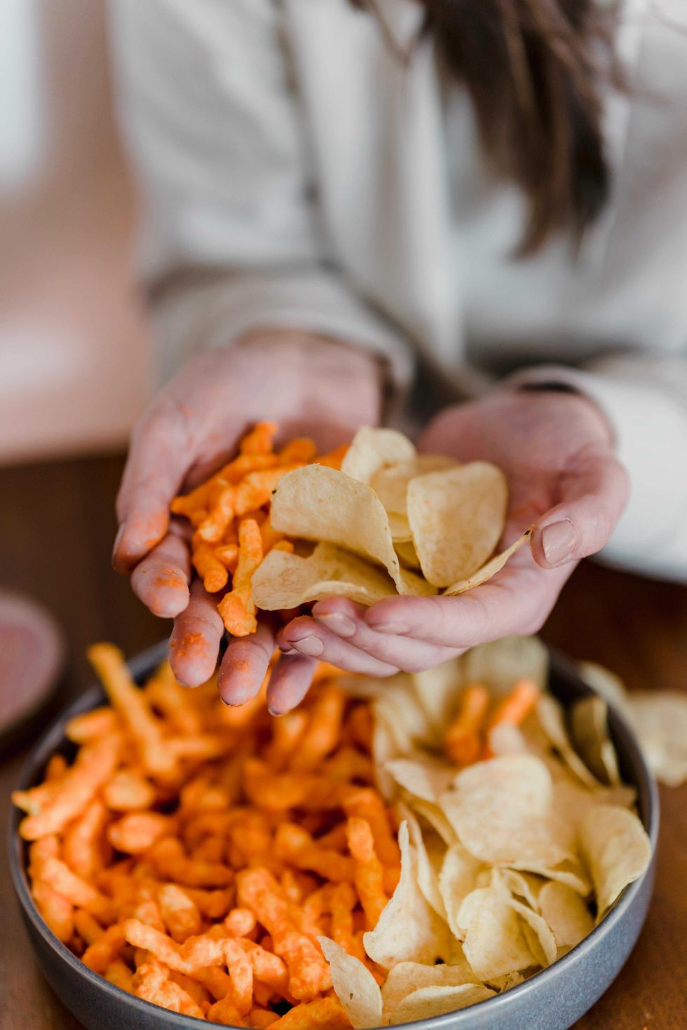 Hands holding cheese puffs and potato chips over a bowl.