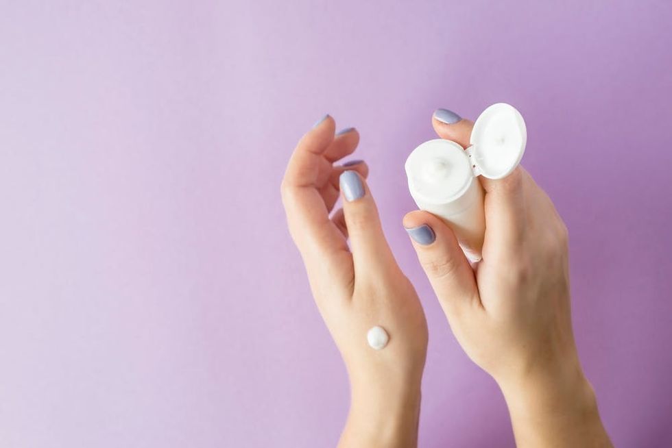 Hands with purple fingernails holding hand cream in front of a purple background.