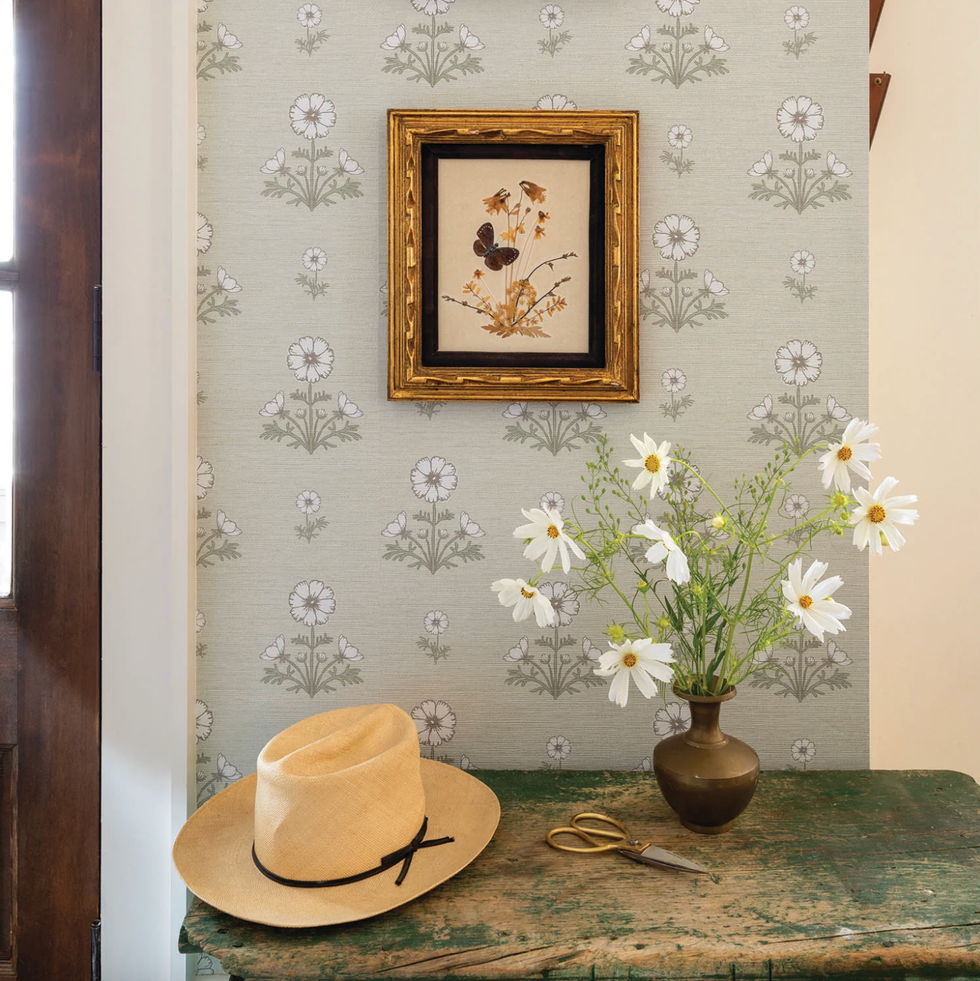 Hat, flowers, and scissors on table with framed butterfly art on floral wallpaper.