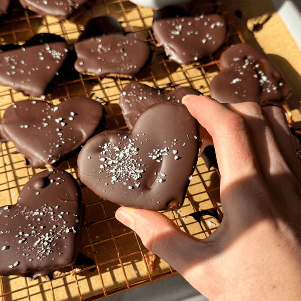Heart-shaped chocolate cookies with sprinkles on cooling rack; hand holding one.