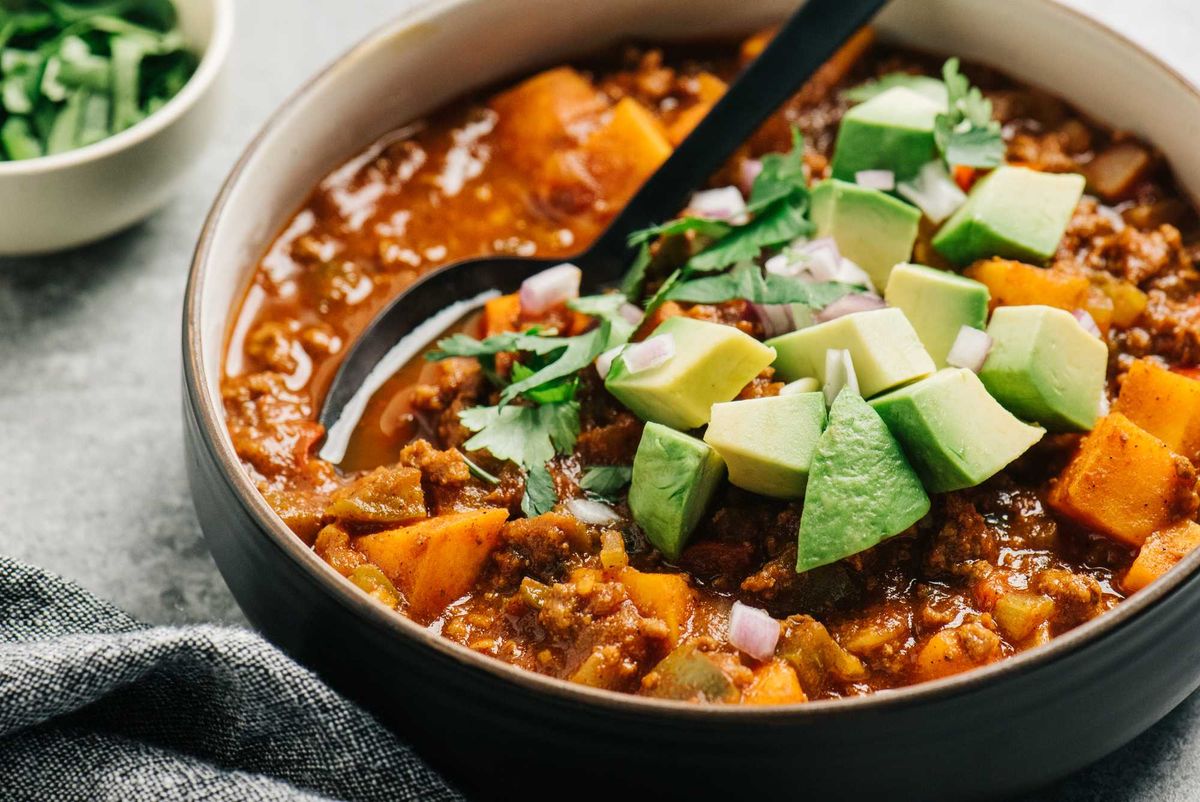Hearty chili with avocado, cilantro, and diced onions in a bowl with a spoon.