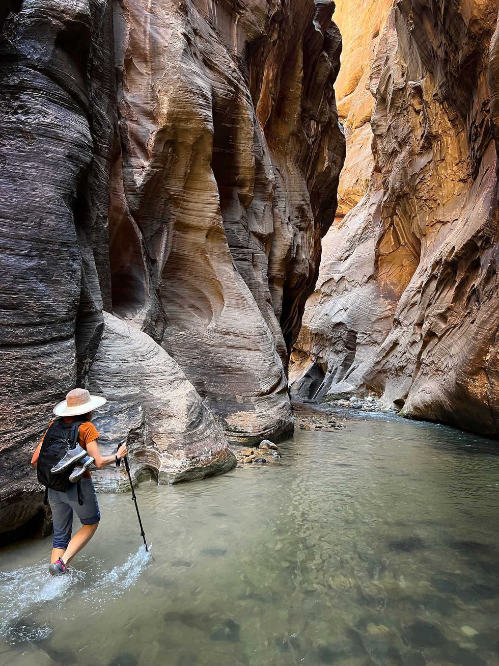 Hiker wades through shallow water in a narrow, scenic canyon with towering rock walls.