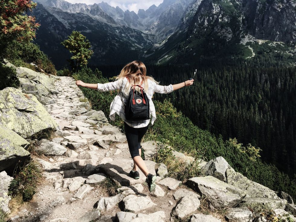 Hiker with a backpack walking on a rocky mountain trail, lush forest and peaks in the background.