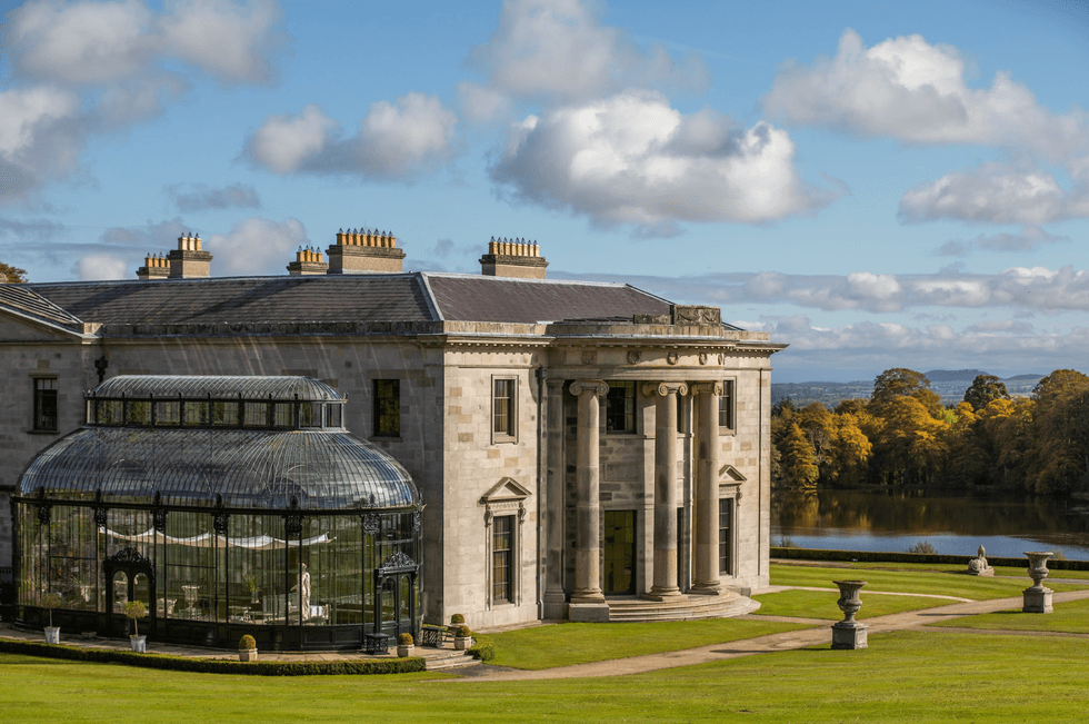 Historic mansion with glass conservatory, set against a lake and autumn trees under a blue sky.