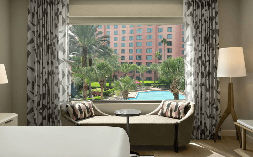Hotel room with a pool and palm tree view through a large window.