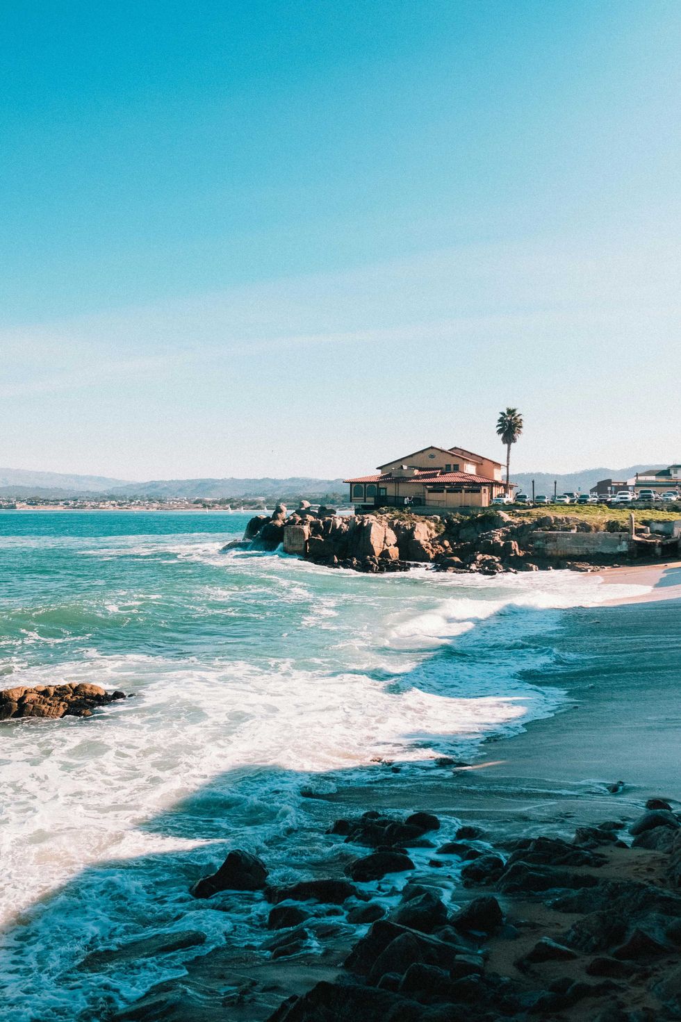 House on a rocky seaside cliff with waves crashing below, under a clear blue sky.