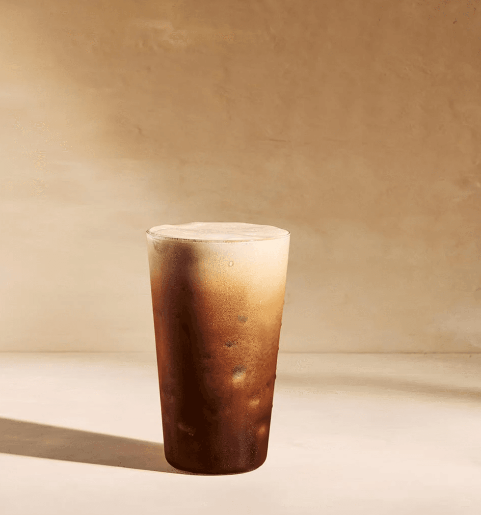 Iced coffee with frothy top in a clear glass, against a beige background.