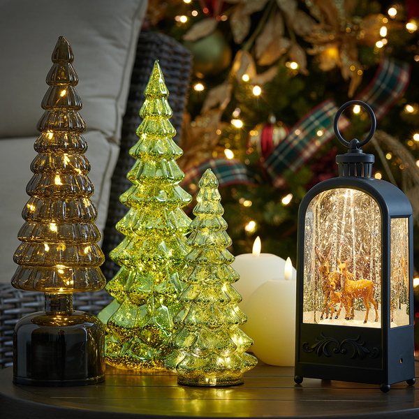 Illuminated glass trees, lantern with deer, and candles by a lit Christmas tree.