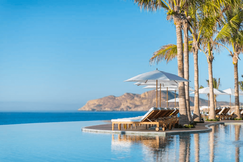 Infinity pool with lounge chairs, umbrellas, and palm trees by the ocean.