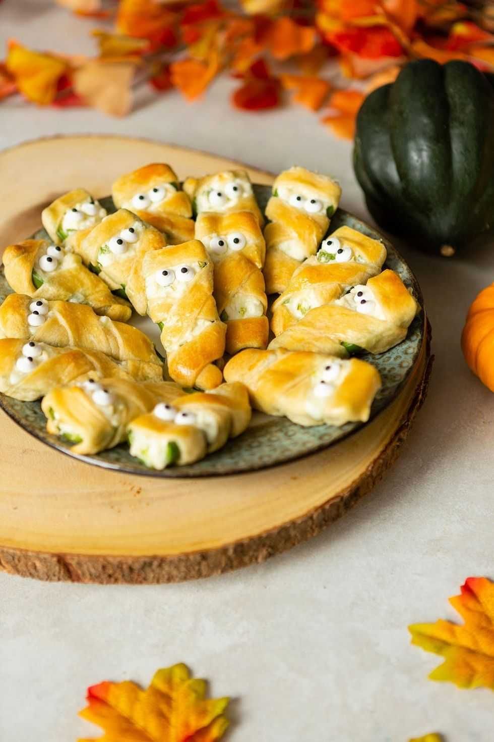 Jalape\u00f1o popper mummies on a plate with autumn leaves and gourds.