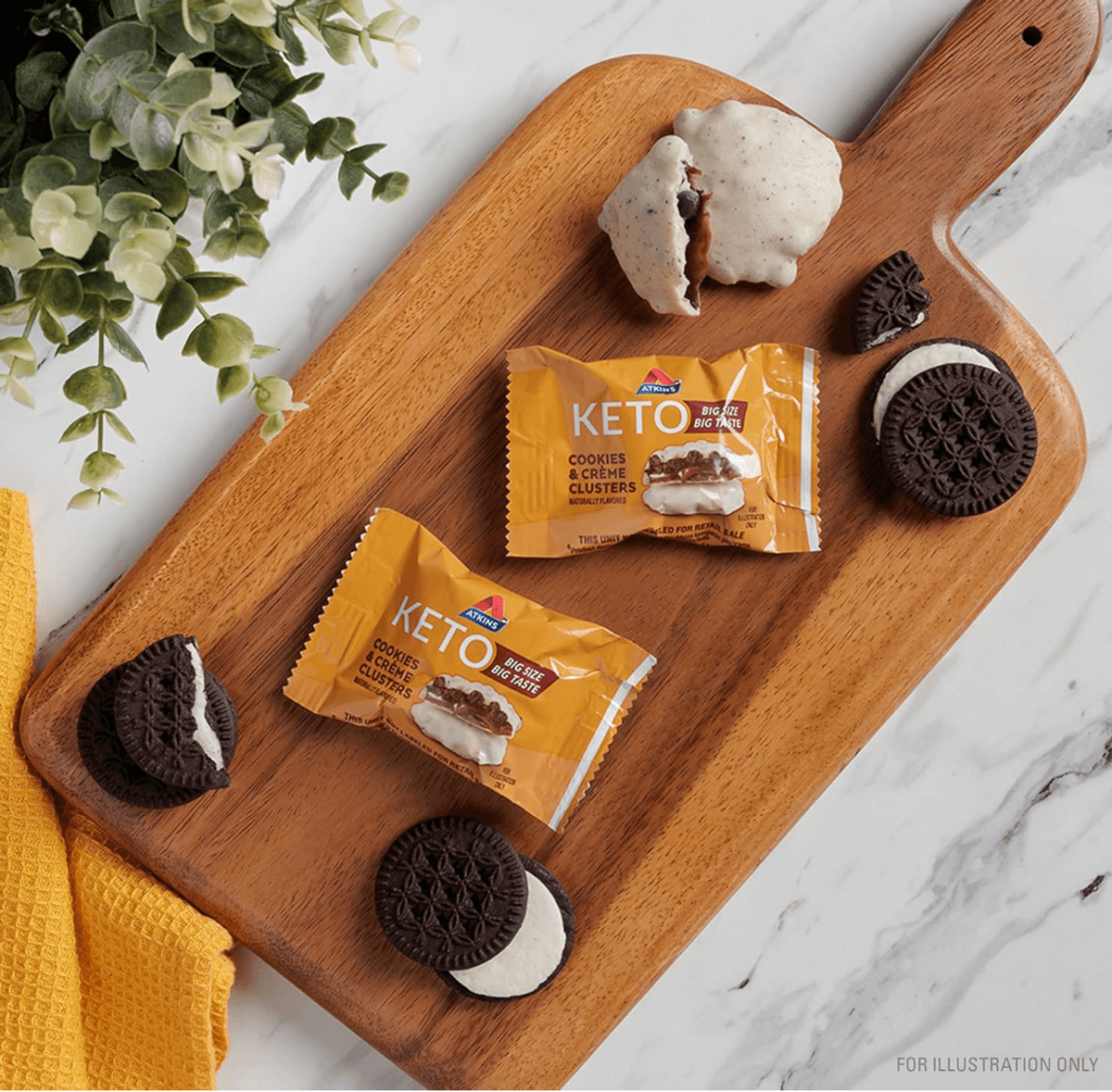 Keto cookie packs and cookies on a wooden board, near a plant and yellow cloth.
