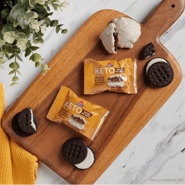 Keto cookie packs and cookies on a wooden board, near a plant and yellow cloth.