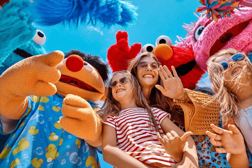 Kids and adults smiling with colorful mascots under a bright blue sky.
