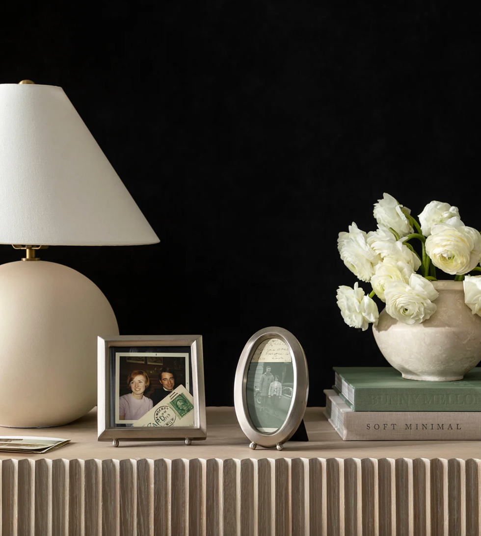 Lamp, framed photos, white flowers, and stacked books on a textured wooden table.