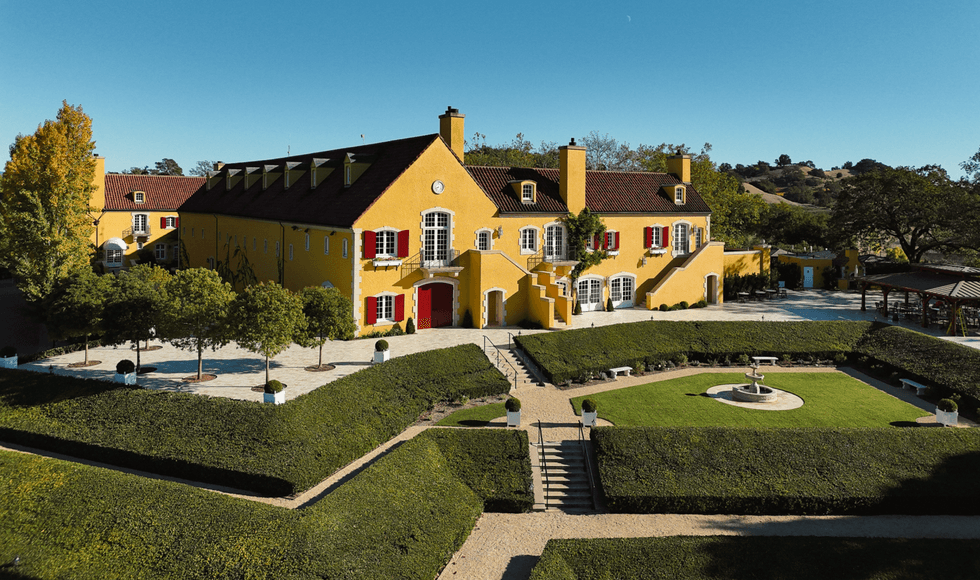 Large yellow chateau with red shutters, surrounded by manicured lawns and hedges.
