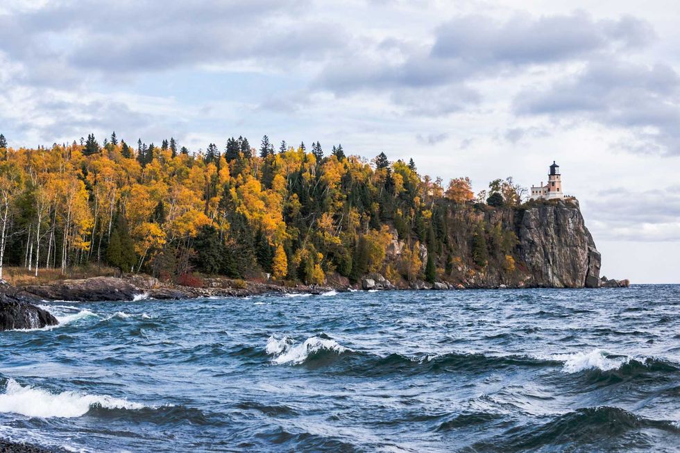 Lighthouse atop a cliff surrounded by autumn trees, overlooking a wavy sea under a cloudy sky.