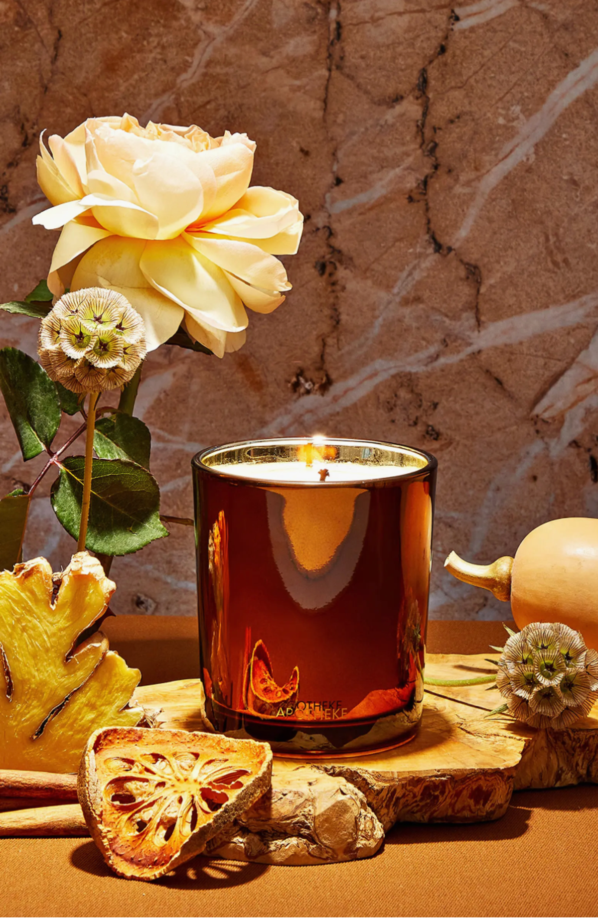 Lit candle in brown holder with flowers and dried fruit on a marble background.