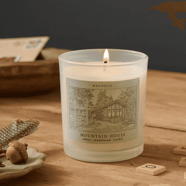 Lit candle labeled "Mountain House" on a wooden table with acorn and feather nearby.