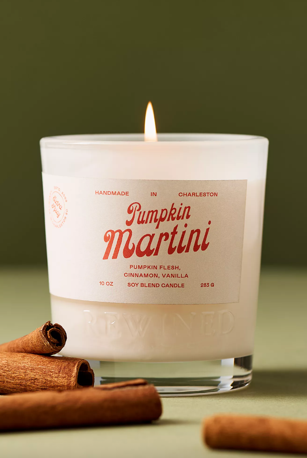 Lit "Pumpkin Martini" candle with cinnamon sticks against olive background.