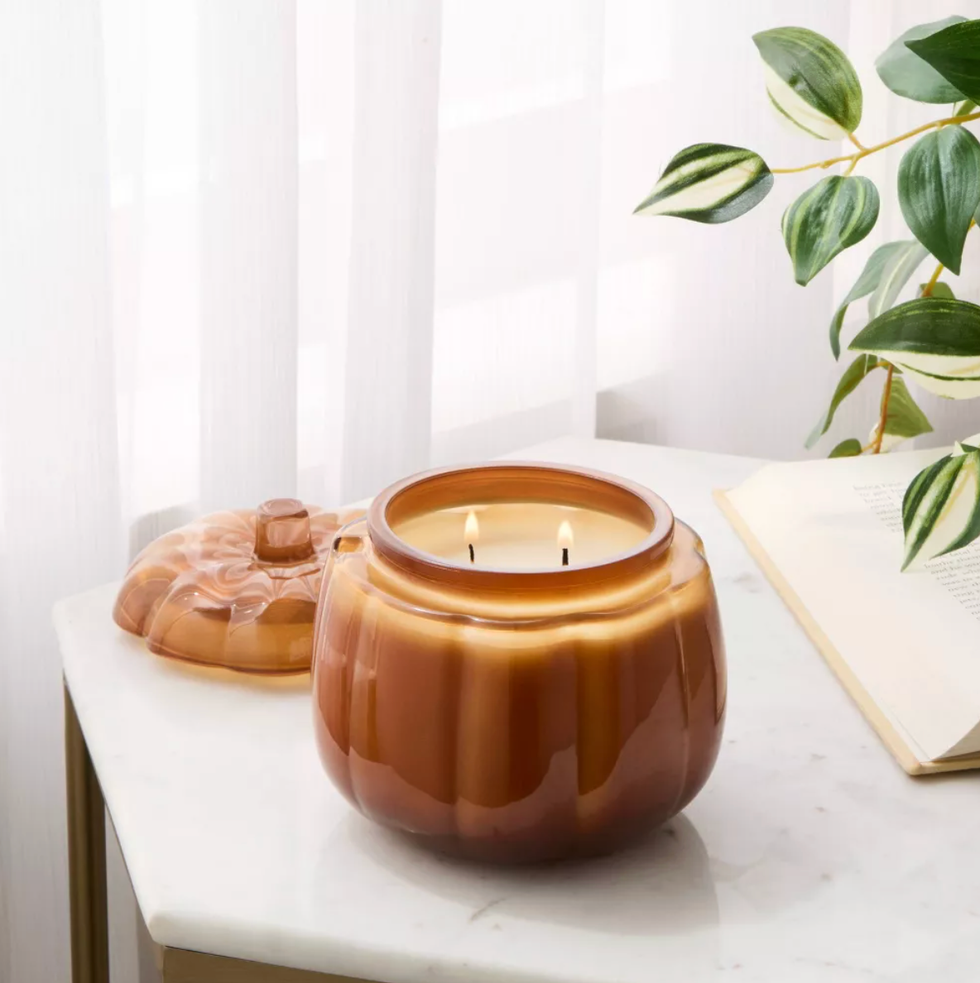 Lit pumpkin-shaped candle on a table with a plant and open book nearby.