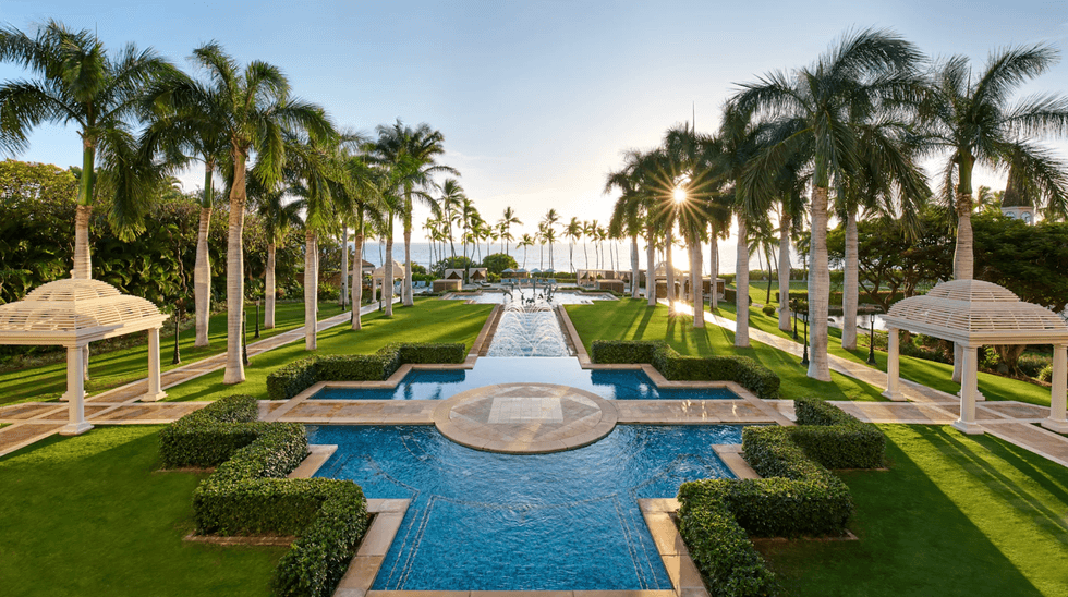 Lush garden with a central fountain, flanked by palm trees and gazebos, at sunset.