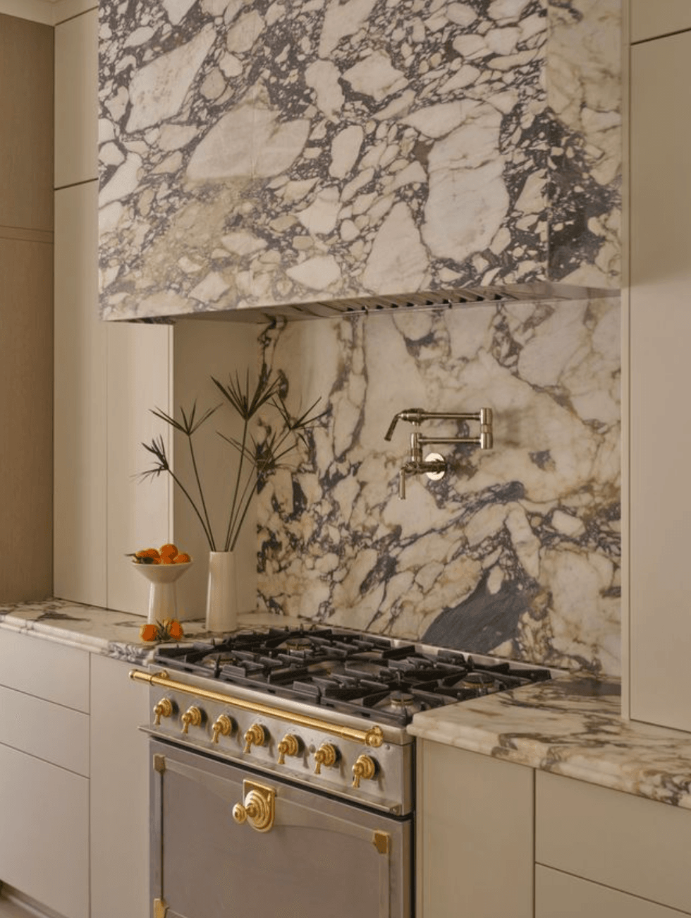 Luxurious kitchen with marble backsplash, gold-accented stove, and fruit bowl on counter.