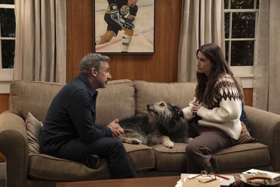 Man and woman seated on a couch with a dog between them, talking in a cozy living room.