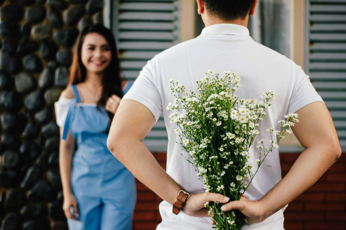 Man hiding daisies behind back, facing a smiling woman outdoors.