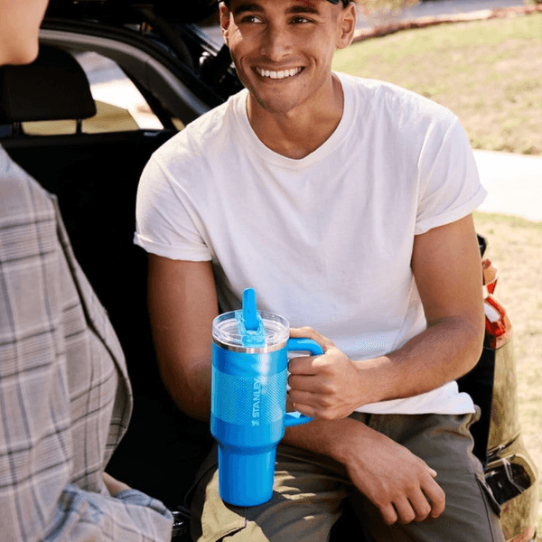 Man in a white T-shirt holding a large blue Stanley cup, sitting in an open car trunk.