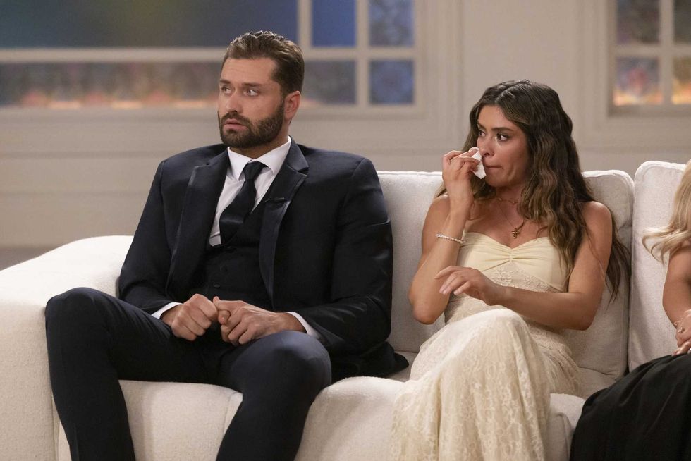 Man in suit and woman in white dress sitting on couch, she wipes her face with tissue.