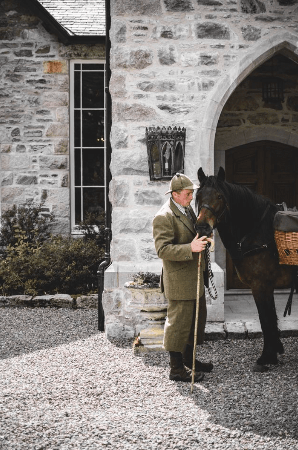 Man in tweed suit with horse outside stone building.